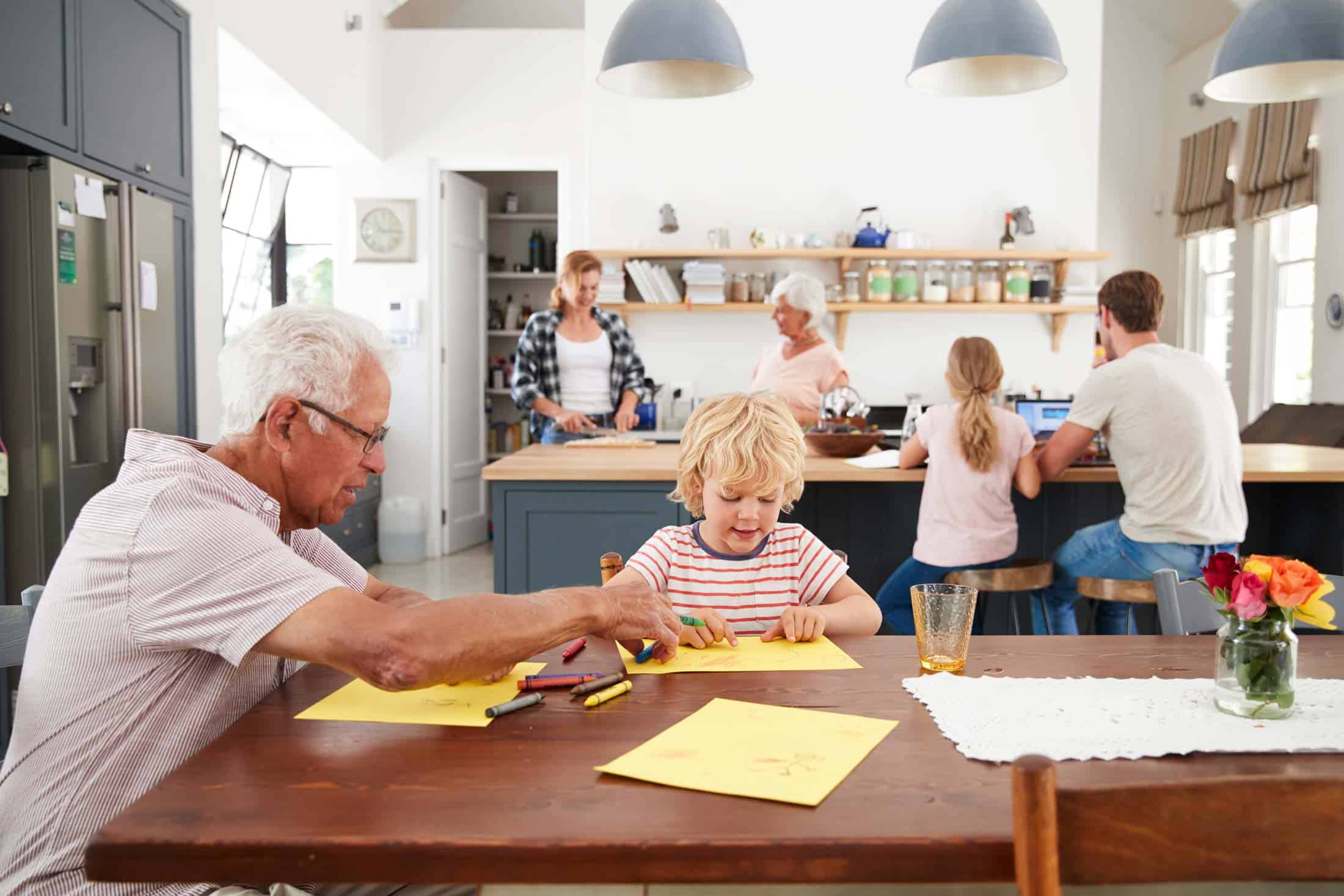 A picture of a family in their kitchen. One older man is helping a small child color a picture on the dining table.